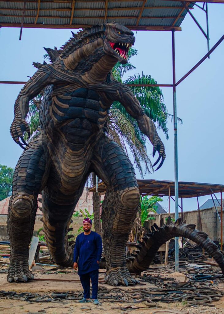 Toyeeb Ajayi standing beneath a towering Godzilla sculpture assembled from strips and pieces of used tyres.