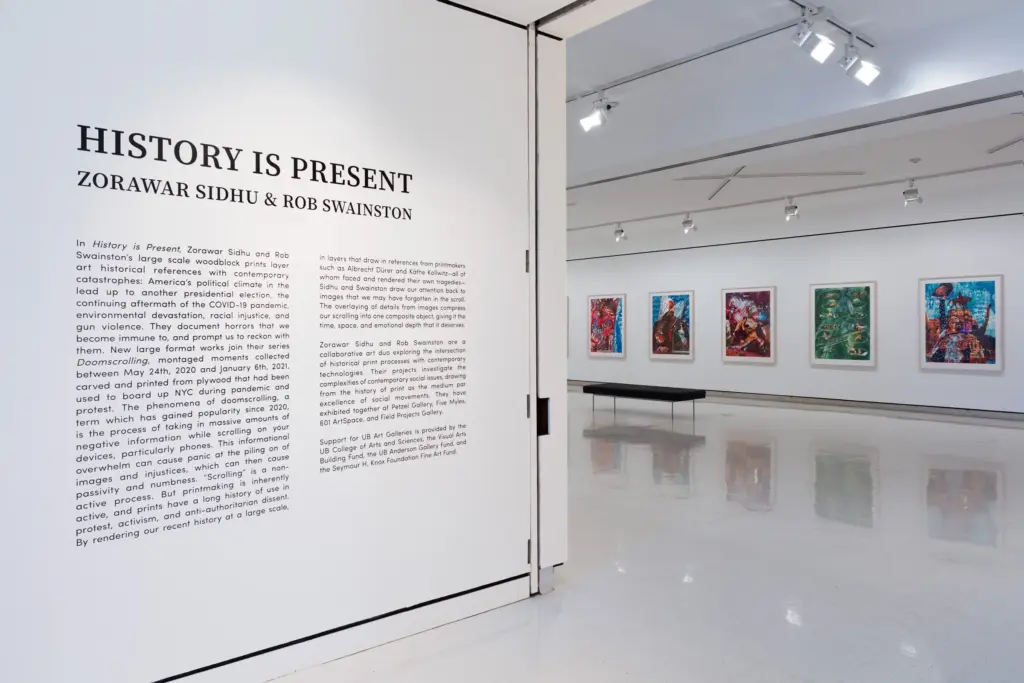 White gallery entrance with wall text reading History Is Present and a row of framed multicolor prints.
